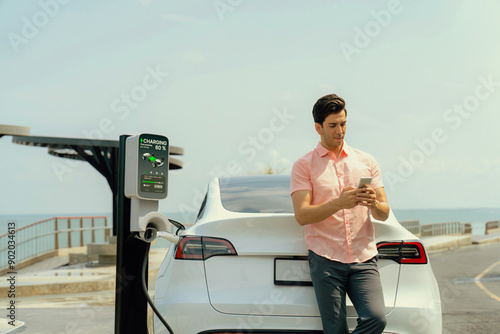 Holiday road trip vacation traveling by the seashore road with electric car, man recharge EV car with green and clean energy while using smartphone. Seascaoe travel with eco-friendly concept.Perpetual photo