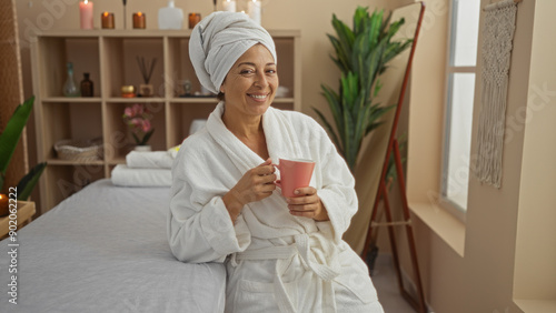 Hispanic woman enjoying a relaxing moment with a mug in a serene spa room adorned with candles and plants.