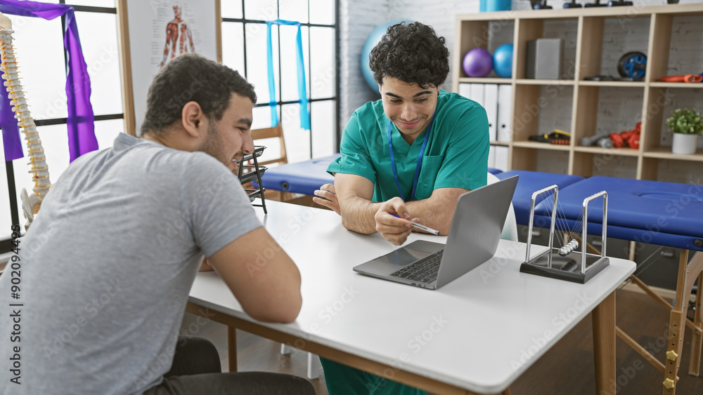 Obraz premium A male therapist consults with a male patient in a modern physiotherapy clinic, discussing treatment over a laptop.
