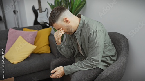Fotografie A distressed man sits on a sofa in a modern apartment, face in hand, showcasing worry or headache in a relatable domestic setting