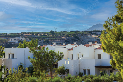 Beautiful view of white two-story hotel buildings with tropical garden plants and mountain pines, set against mountain backdrop on Crete.