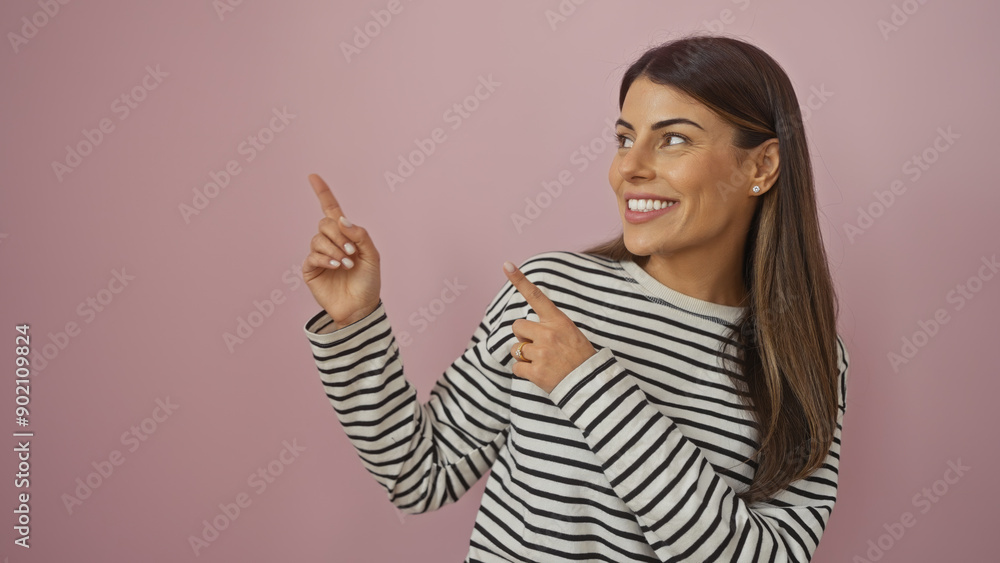 A cheerful young hispanic woman in a striped shirt poses against a pink background, pointing upwards with both hands. A cheerful young hispanic woman in a striped shirt poses against a pink background, pointing upwards with both hands.