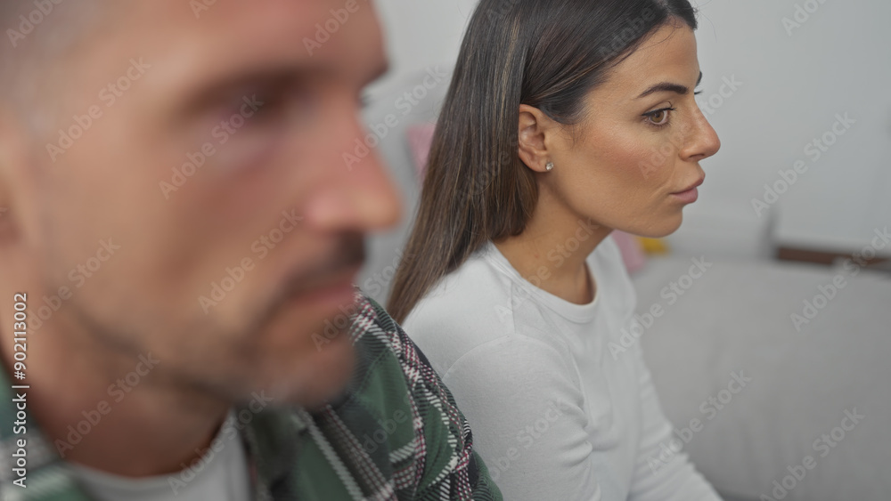Fototapeta premium A contemplative man and woman sitting apart on a sofa in a well-lit living room, symbolizing relationship issues.