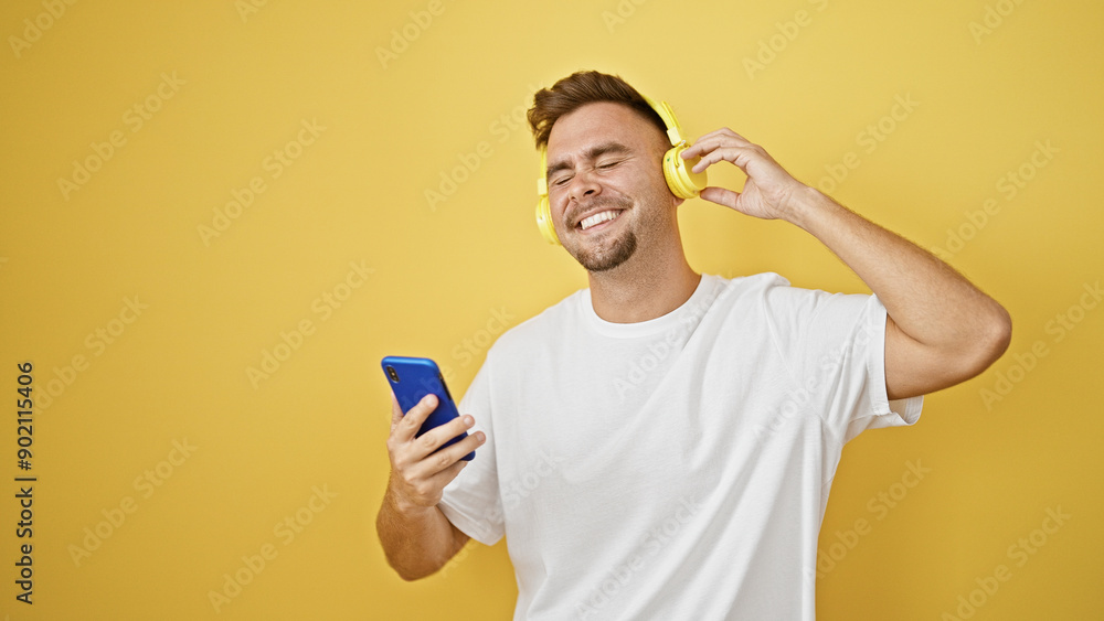Smiling young man with beard enjoying music on yellow headphones against isolated yellow background, showcasing positive emotion. Smiling young man with beard enjoying music on yellow headphones against isolated yellow background, showcasing positive emotion.