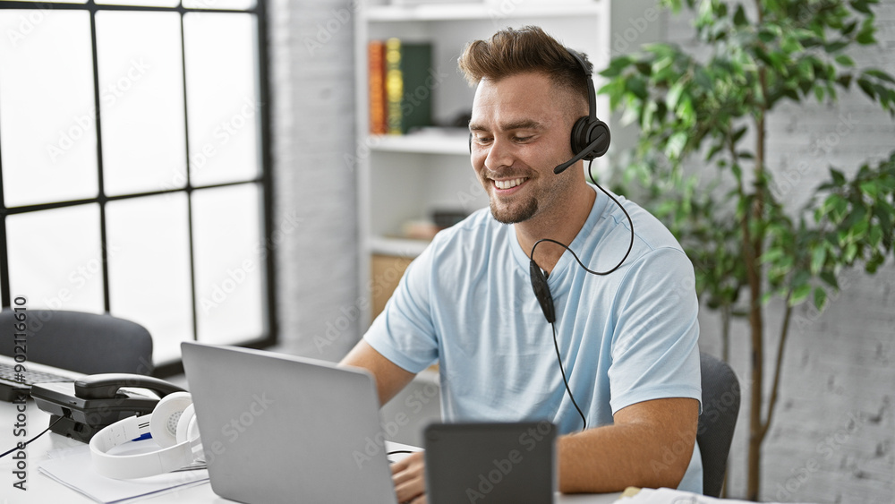A cheerful young man with a headset working on a laptop in a bright office space A cheerful young man with a headset working on a laptop in a bright office space