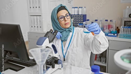 A mature woman scientist in hijab examines a test tube in a laboratory setting, conveying a sense of professionalism and expertise.