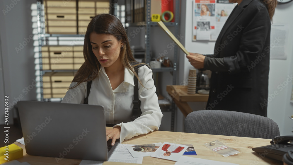 Two women investigators analyze evidence at a police department with a laptop, documents, and crime photos on desk. Two women investigators analyze evidence at a police department with a laptop, documents, and crime photos on desk.