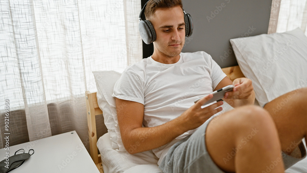 Fototapeta premium A young man relaxing in bed with headphones and a smartphone in a cozy bedroom setting