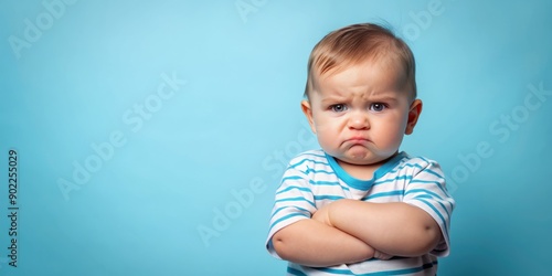 Adorable infant boy with a furrowed brow and crossed arms conveys displeasure against a bright blue studio backdrop with a subtle gradient effect.