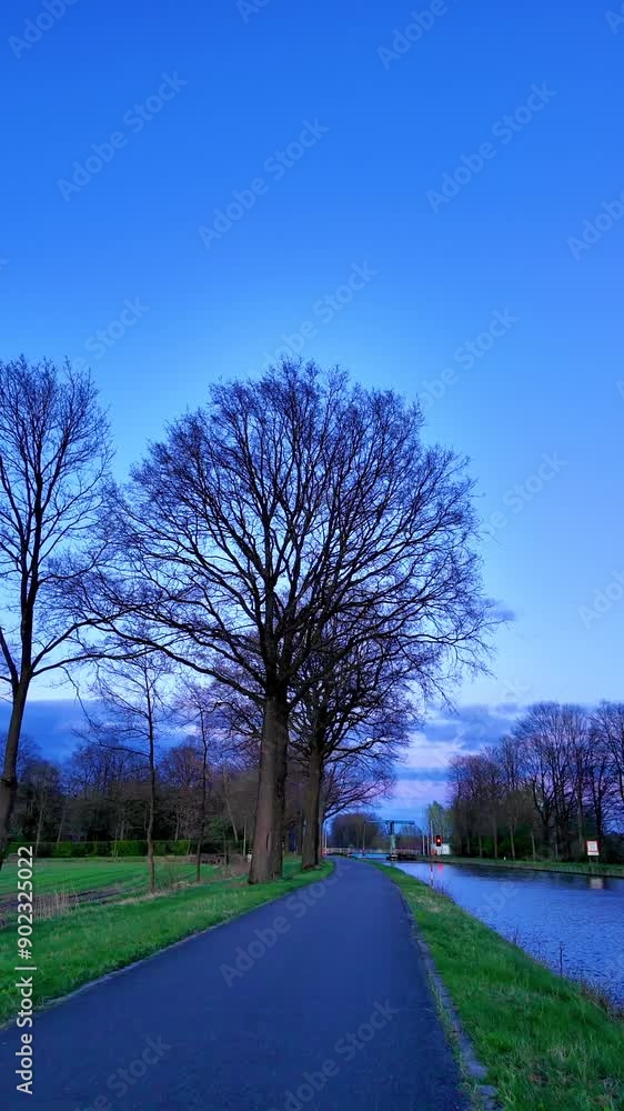 A tranquil evening view featuring a pathway, bare trees, and a calm river under a beautiful blue sky in the countryside A tranquil evening view featuring a pathway, bare trees, and a calm river under a beautiful blue sky in the countryside