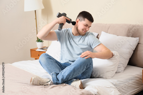 Young man massaging his neck with percussive massager in bedroom