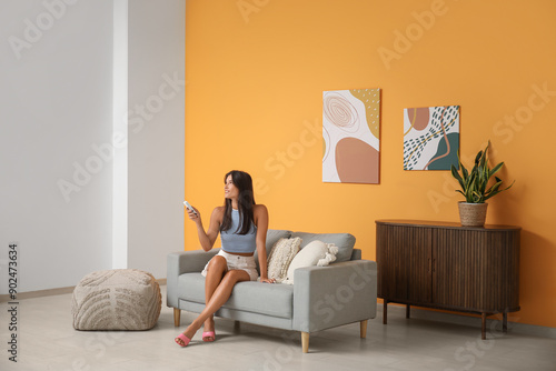 Young woman with air conditioner remote control sitting on sofa near orange wall in living room