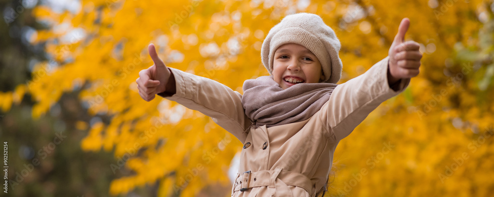 Happy caucasian girl in a beige coat and beret walks in the park in autumn. Schoolgirl showing thumbs up. 