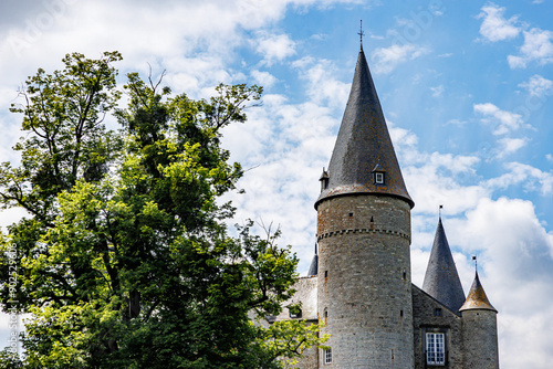 Towers with conical roofs and stone walls of Veves Castle, huge tree with green leaves, sunny day with blue sky and white clouds in Houyet, Namur province, Wallonia, Belgium photo