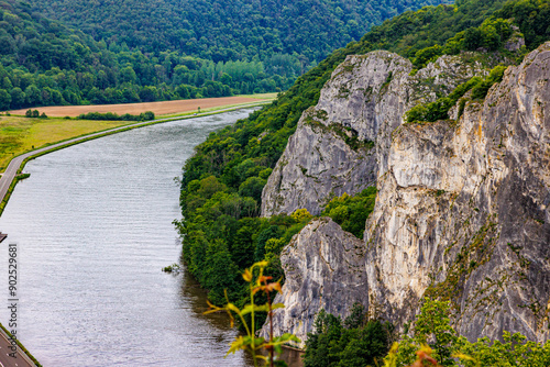 Meuse River and rock formation known as Freyr Rocks, among meadow, hills with abundant green leafy trees, irregular rock wall famous place for climbing, cloudy summer day in Wallonia, Belgium