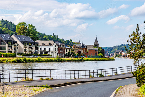 Rural road towards city of Dinant next to Meuse river, buildings, houses with hill covered with green leafy trees in background, sunny summer day with blue sky in Namur province, Wallonia, Belgium photo