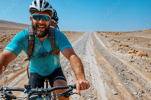 Bearded cyclist grins with joy while riding mountain bike on remote desert road, wearing turquoise jersey and helmet, adventure and endurance in harsh terrain photo