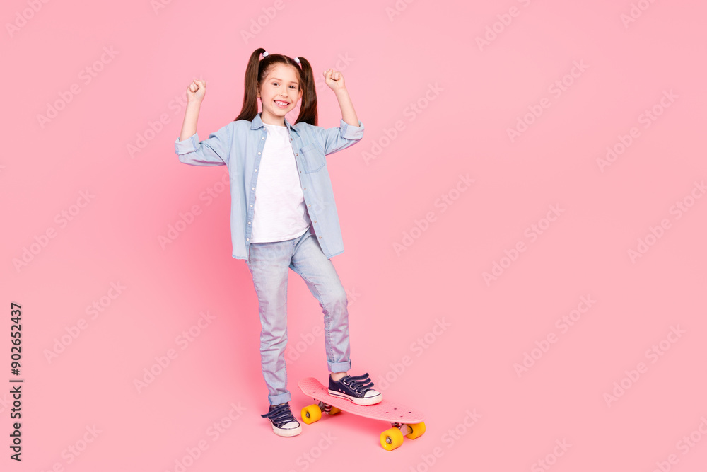 Full size photo of positive small girl dressed denim shirt pants stand on skateboard clenching fists isolated on pink color background Full size photo of positive small girl dressed denim shirt pants stand on skateboard clenching fists isolated on pink color background