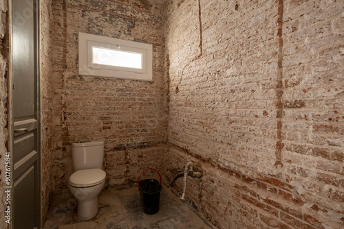 An indoor bathroom space, featuring exposed brick walls, a toilet, small window, and construction materials, illustrating the room’s renovation phase.