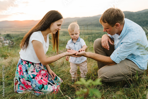 Mother, father feeds baby boy blackberries and on a picnic in mountains. Funny face closeup. Mom, dad play with son in sunset. Happy family holiday on summer day. Parents with child. Side view. © Serhii