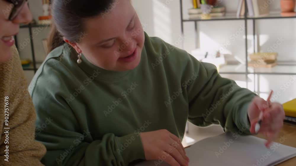 Girl with Down syndrome writing down notes in copybook while studying at home during private lesson with female tutor Girl with Down syndrome writing down notes in copybook while studying at home during private lesson with female tutor