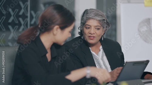 Indian older aged boss women use tablet touch pad look screen talk new deal plan share ideas with teen girl indoor job office young adult expert female old elder lady staff do work day task work place