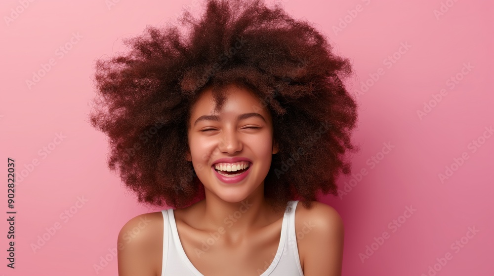 A woman with curly hair is smiling and laughing. She is wearing a white tank top and is standing in front of a pink background A woman with curly hair is smiling and laughing. She is wearing a white tank top and is standing in front of a pink background