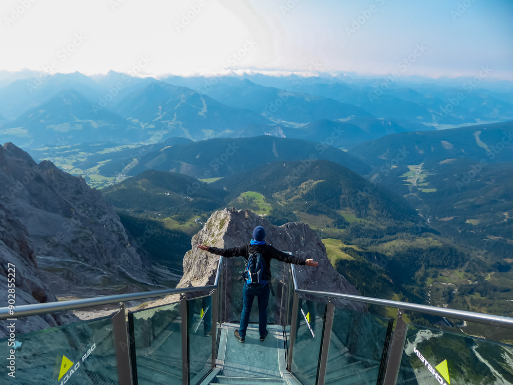 Young man with backpack stands on observation deck of skywalk rope bridge at Dachstein Mountains, Northern Limestone Alps, Styria, Austria. Looking at mountain ranges of Austrian Alps. Peace of mind