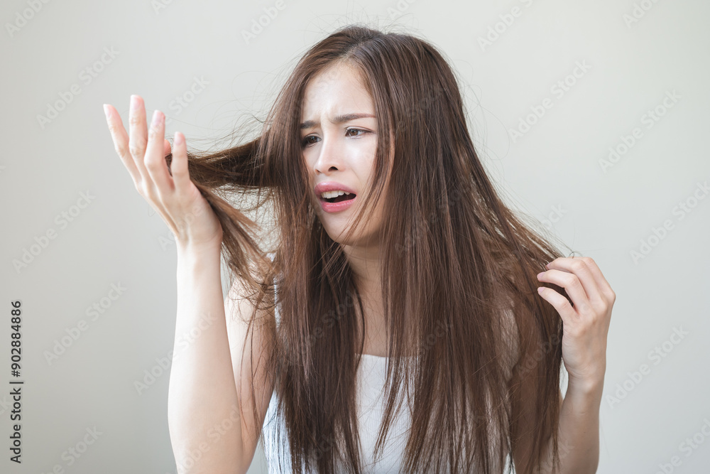 Fototapeta premium Damaged Hair, frustrated asian young woman, girl hand in holding splitting ends, messy unbrushed dry hair with face shock, long disheveled hair, health care of beauty. Portrait isolated on background.