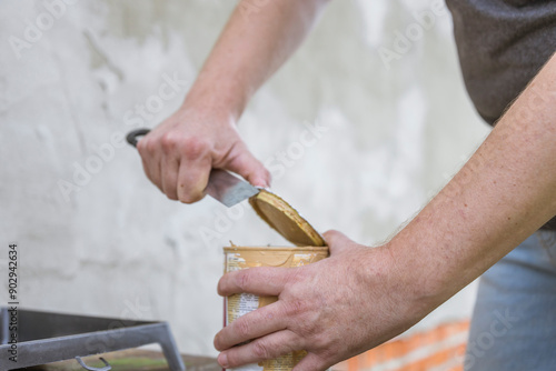 A man's hands use a utility knife to carefully pry open a can of paint, likely for a home renovation project.