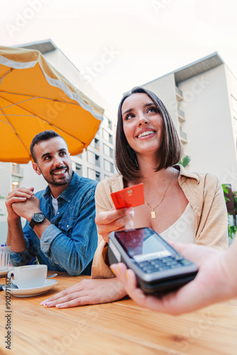 Vertical couple paying the bill of the coffee shop using a contactless creditcard payment. Young adult customer woman doing a purchase on a restaurant to the cashier with a debit card. People spending