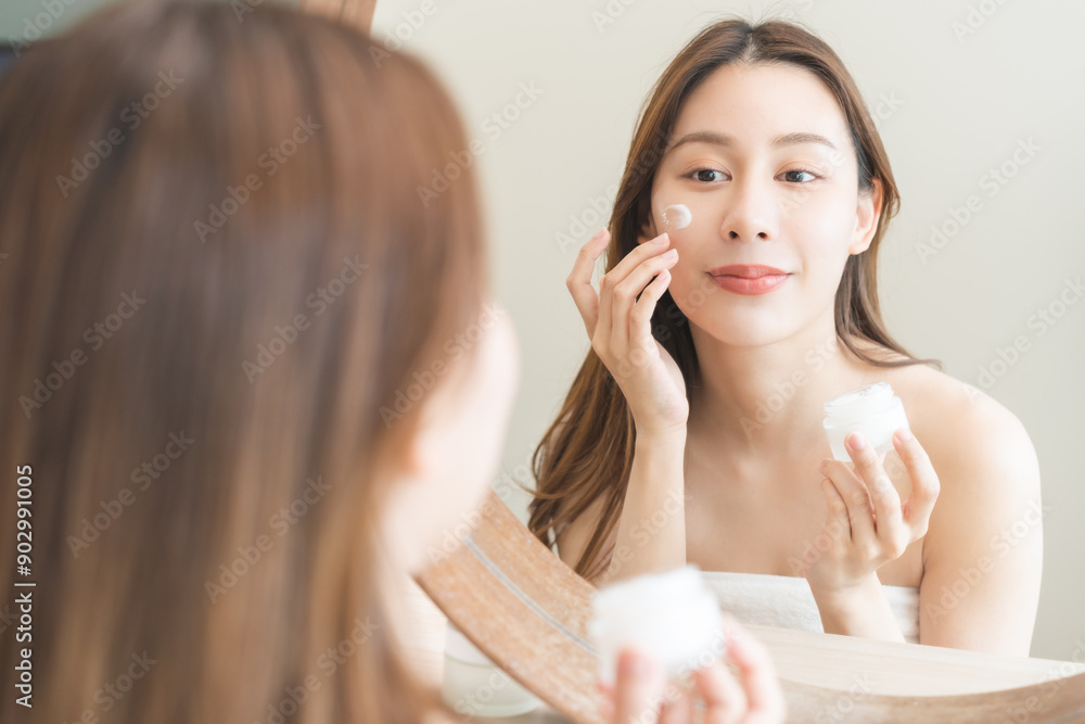Facial beauty skin care, smile of pretty asian young woman in bathrobe looking at mirror, hand applying moisturizer lotion on her face, holding jar of skin cream before makeup cosmetic routine at home Facial beauty skin care, smile of pretty asian young woman in bathrobe looking at mirror, hand applying moisturizer lotion on her face, holding jar of skin cream before makeup cosmetic routine at home