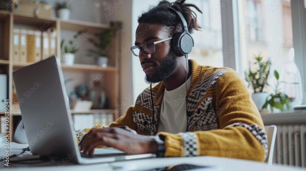A young man wearing headphones focuses intently on his laptop in a bright, stylish home office filled with plants. A young man wearing headphones focuses intently on his laptop in a bright, stylish home office filled with plants.