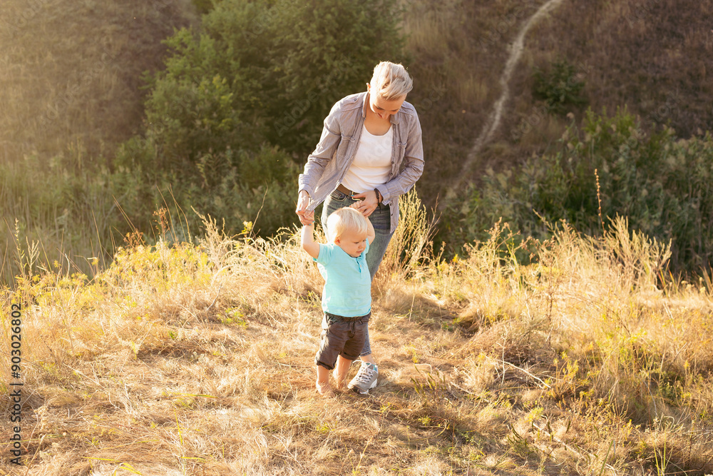 Little boy child takes his first steps and mother holds his hands in summer on grass with sun rays on sunset Little boy child takes his first steps and mother holds his hands in summer on grass with sun rays on sunset