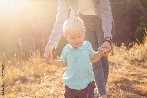 Little baby boy child takes his first steps and mother holds his hands in summer on grass with sun rays on sunset