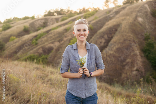 Portrait of smiling young woman with short haircut hairstyle wearing blue shirt in nature outdoors on sunset near green hills