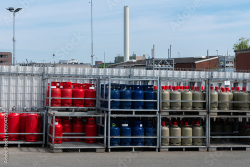 Quadro su tela Different colored gas cylinders are organized in a storage facility under clear skies, with industrial structures in the background