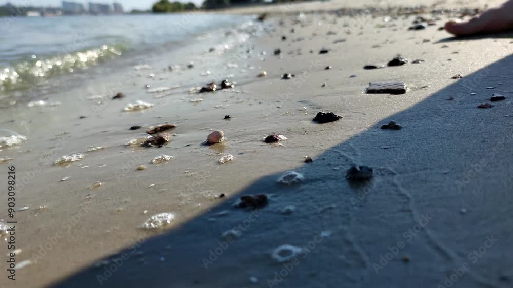 Close-up side view of sand and stones on river beach in a sunny day. Blurred walking barefoot male person in the background. Product placement. Copy space. Selective focus. Leisure activity theme. Close-up side view of sand and stones on river beach in a sunny day. Blurred walking barefoot male person in the background. Product placement. Copy space. Selective focus. Leisure activity theme.