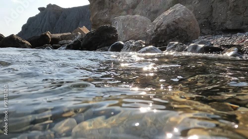 Glare of the sun in the transparent sea, small waves and ripples, behind the mountains and a wild rocky beach, shooting in shallow water