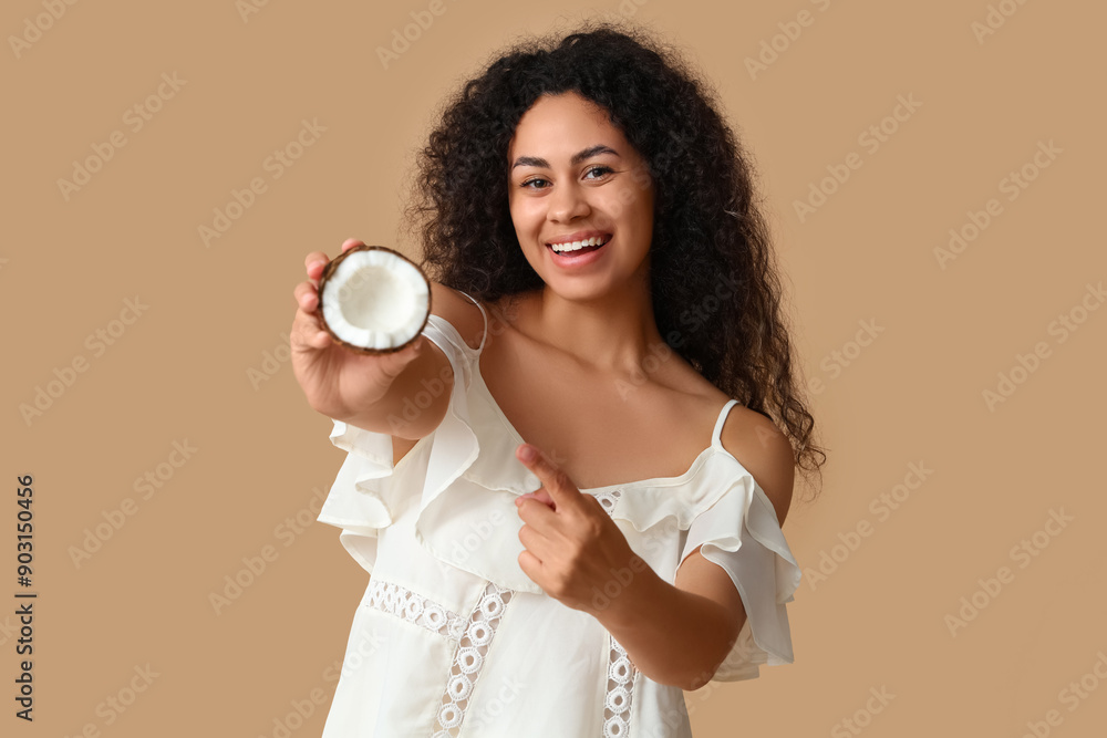 Beautiful young African-American woman pointing at half of fresh coconut on brown background Beautiful young African-American woman pointing at half of fresh coconut on brown background
