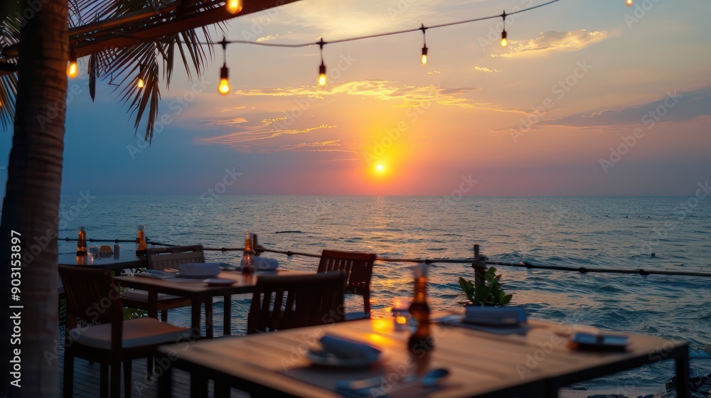 Fototapeta premium A beachfront restaurant with tables and chairs overlooking the ocean. The sun is setting, creating a warm and inviting atmosphere