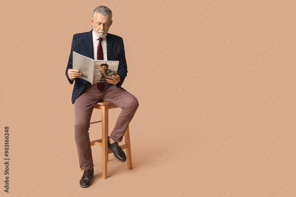 Fototapeta premium Handsome mature man in stylish formal suit with tie and magazine sitting on chair against beige background