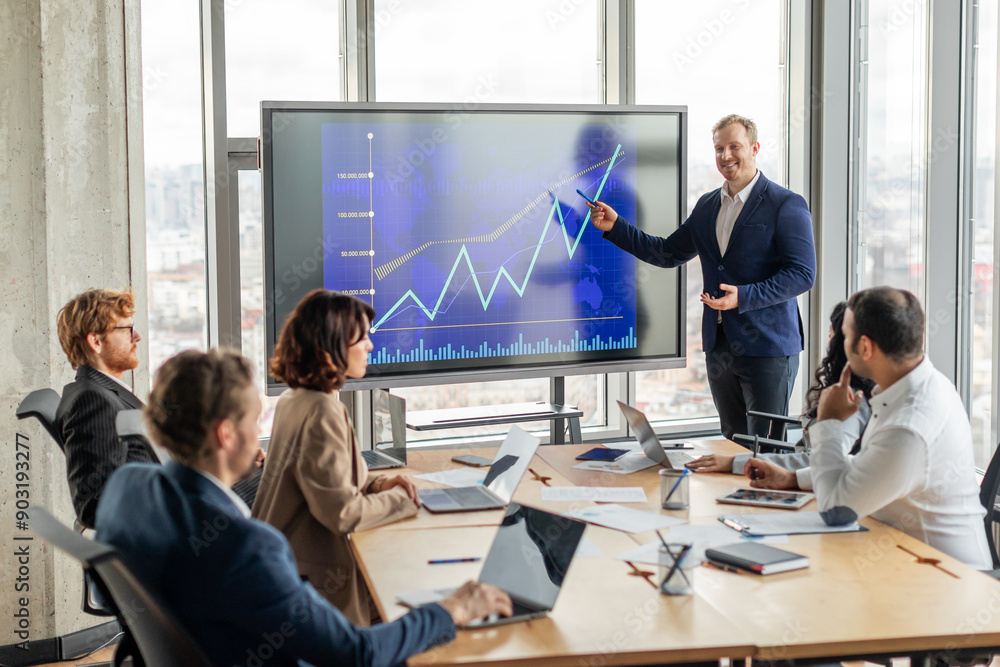 © Prostock-studio - A businessman stands in front of a large screen displaying a blue line graph that is trending upward, pointing to screen with a pen as he explains positive results to a group of attentive colleagues