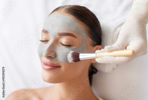 Beautician at spa salon applying grey natural clay face mask to young woman, lady enjoying wellness and skin care treatments, closeup photo