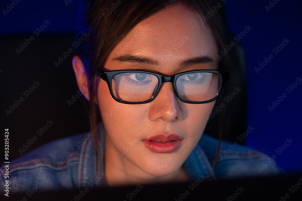 Fototapeta premium Portrait young beautiful Asian creative with glasses of serious face looking on pc to search project creator social media online, planning strategy creator at neon modern office at night. Stratagem.