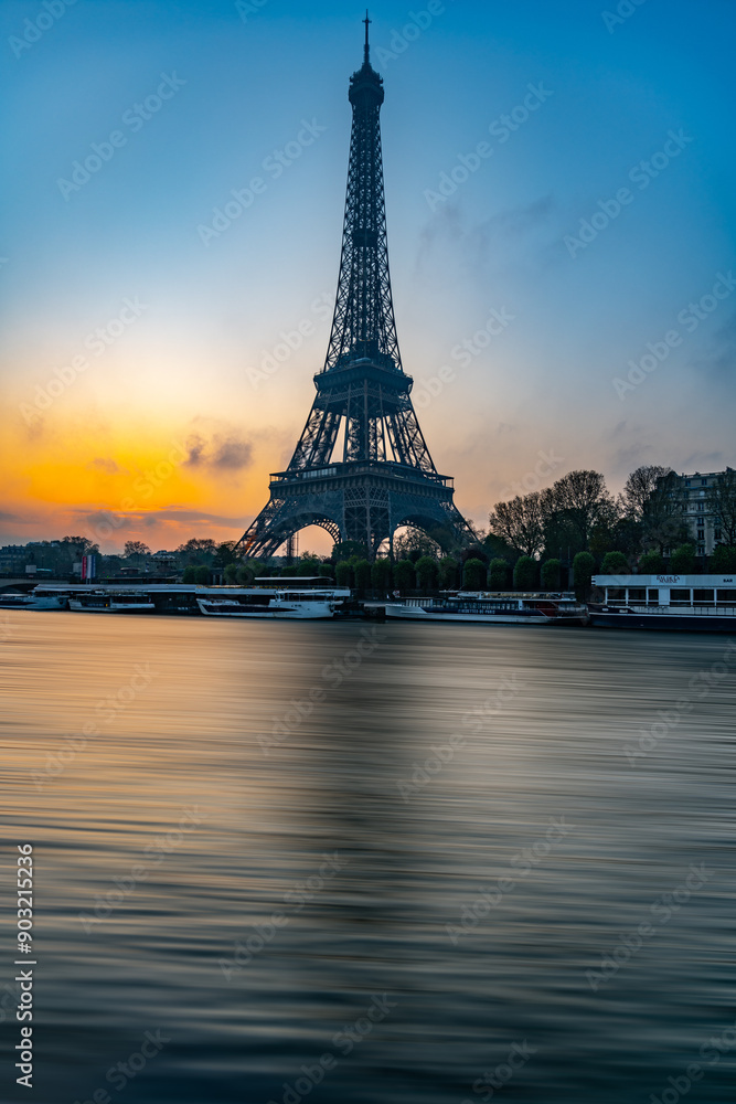 Naklejka premium The Eiffel Tower stands majestically against a colorful morning sky as the sun rises, casting reflections on the calm waters of Seine River nearby.