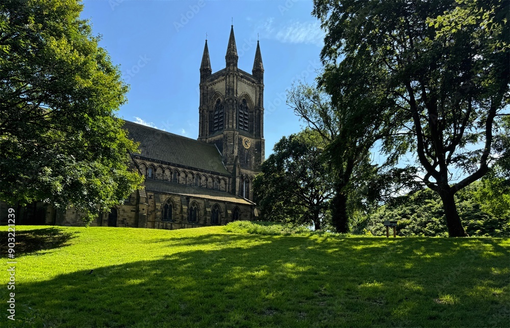 Naklejka premium St Mary's grand Gothic-style church rises majestically among verdant trees, with sunlight filtering through to cast dappled shadows across the grassy lawn in Mirfield, Yorkshire, UK.
