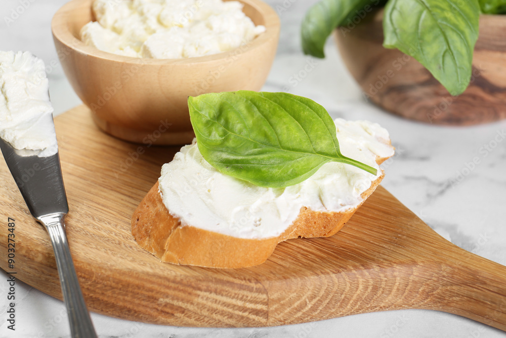 Piece of bread with cream cheese, basil and knife on white marble table Piece of bread with cream cheese, basil and knife on white marble table
