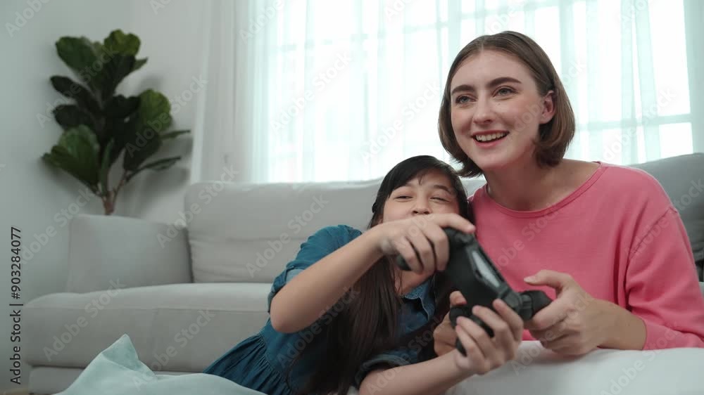 Caucasian mom and daughter sitting and playing game at living room. Happy mother spend time together with smart asian girl while holding joystick ans enjoy playing games. Lifestyle concept. Pedagogy. Caucasian mom and daughter sitting and playing game at living room. Happy mother spend time together with smart asian girl while holding joystick ans enjoy playing games. Lifestyle concept. Pedagogy.
