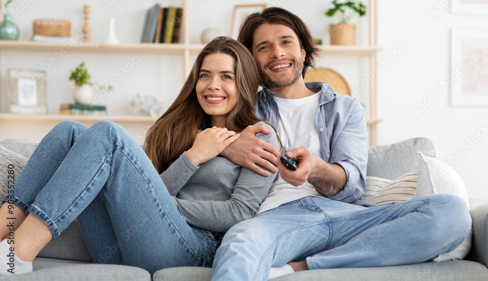 Obraz premium Joyful young couple watching TV together at home, embracing and sitting on couch. Cheerful man and woman switching channels on TV with remote, seeking nice movie for weekend, empty space